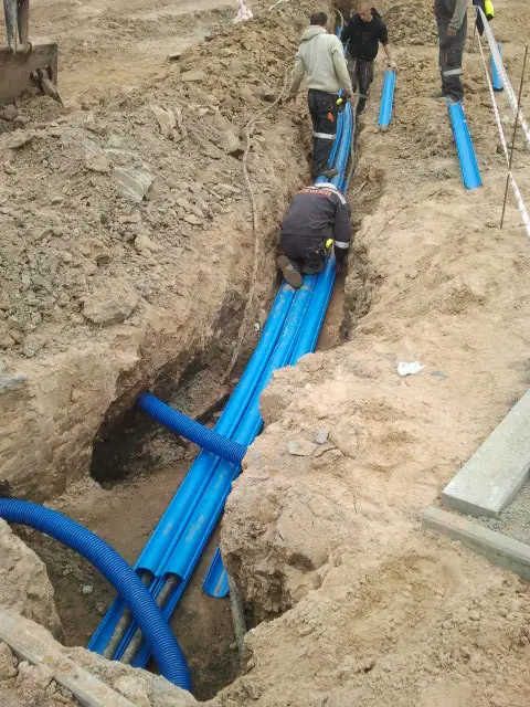 Ekipa układa niebieskie rury instalacyjne w głębokim wykopie oznaczonym tyczkami