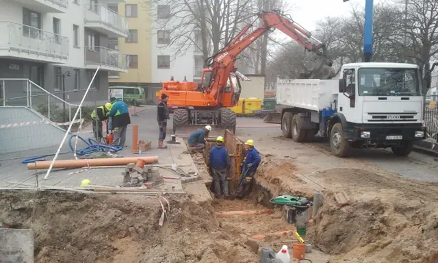 Koparka i ciężarówka pomagają przy układaniu rur w wykopie na terenie budowy osiedla