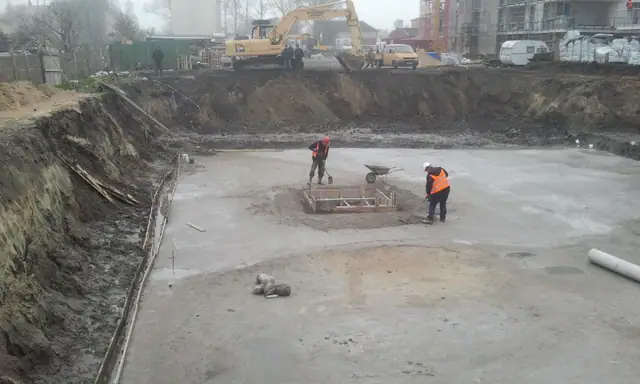 Robotnicy wyrównują podłoże taczką przy niewielkim szalunku w wykopie fundamentowym