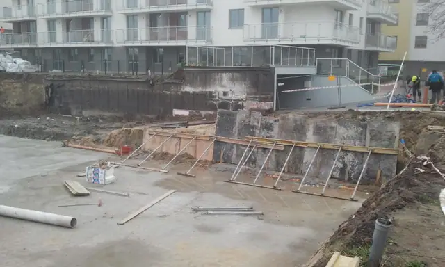 Głęboki wykop z drewnianym szalowaniem ścian tuż obok gotowego bloku mieszkalnego