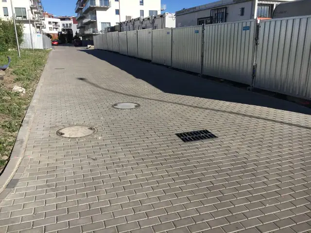 Prosta droga z szarej kostki brukowej wzdłuż blaszanego płotu ze studzienkami