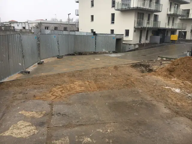 Wyrównany plac betonowy ogrodzony blaszanym płotem przy budynkach mieszkalnych osiedla