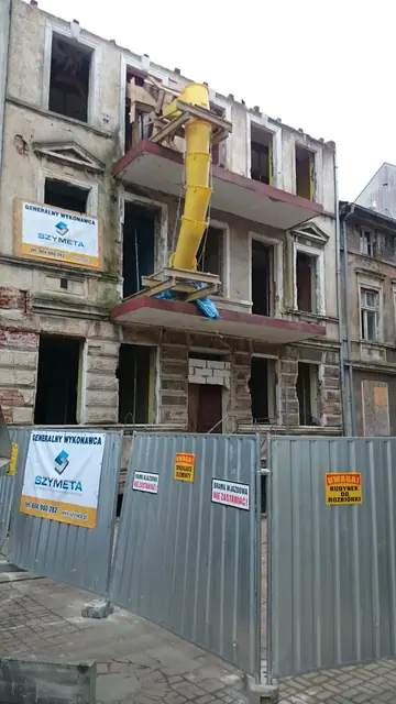 Koparka z młotem wyburzeniowym pracuje przy elewacji kamienicy ogrodzonej płotem budowy