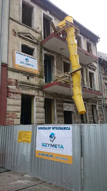 Hydrauliczne ramię wyburzeniowe koparki nad dachem kamienicy, ogrodzenie z banerem SZYMETA