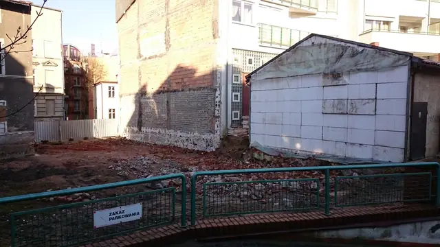 Ogrodzony teren po rozbiórce z zachowanym białym budynkiem gospodarczym w tle