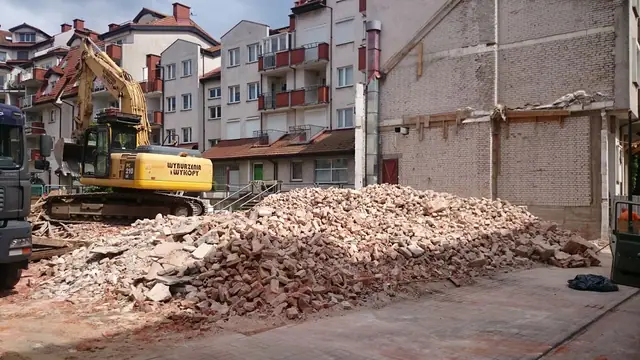 Koparka wyburza ostatnie fragmenty muru na tle wielorodzinnej zabudowy mieszkalnej