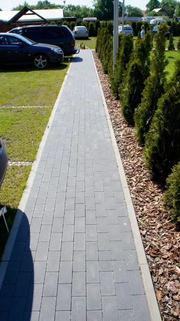 Wybrukowana alejka obsadzona młodymi tujami i rabatami z korą prowadzi do domków