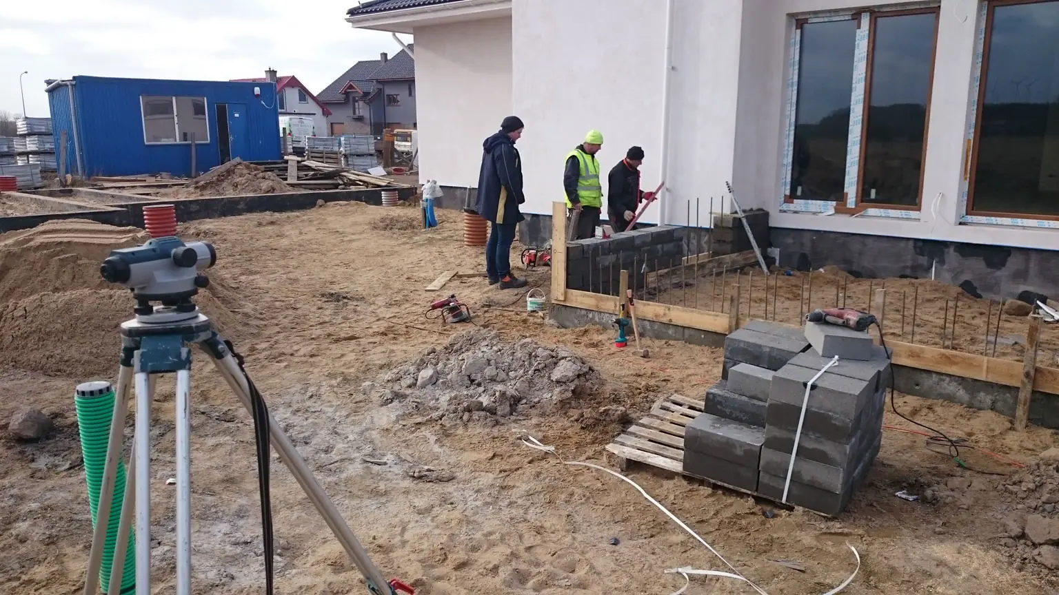 Geodeta z niwelatorem mierzy teren przy murowanym fundamencie ogrodzenia na granicy posesji