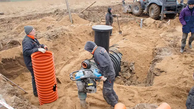 Robotnicy montują pomarańczową rurę drenażową w wykopie obok zagęszczarki gruntu