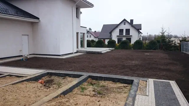 Świeżo obsiana czarną ziemią działka wokół domu, w tle sąsiednie zabudowania