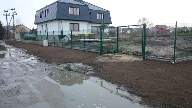 Nowo wybudowany dom za zielonym ogrodzeniem z siatki, błotnisty teren posesji