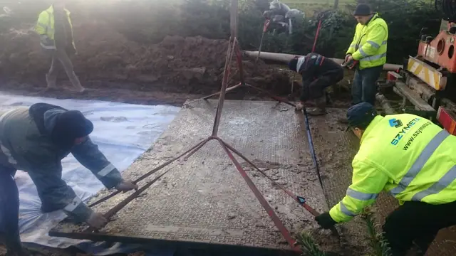 Robotnicy w kamizelkach Szymeta układają maty drogowe na geowłókninie za pomocą dźwigu