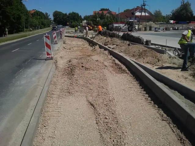 Długi odcinek żwirowej jezdni z nowo ułożonym krawężnikiem, w oddali pracuje ekipa