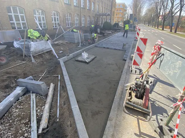 Robotnicy układają płyty chodnikowe przy zabytkowej elewacji, obok leżą elementy betonowe