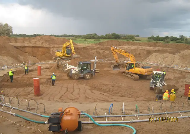 Robotnicy rozprowadzają węże do odwadniania w szerokim wykopie pod fundament turbiny wiatrowej