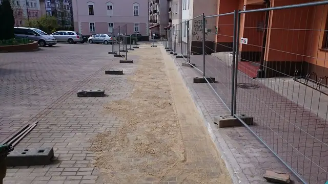 Robotnicy układają kostkę brukową na parkingu ogrodzonym siatką przy kamienicach