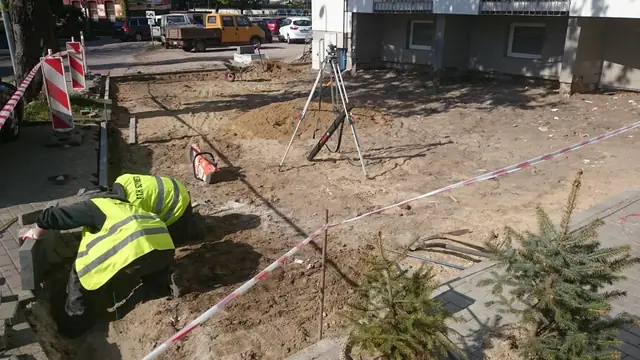 Robotnik wytycza teren pod nawierzchnię przy trójnogu niwelatora obok świerków w donicach