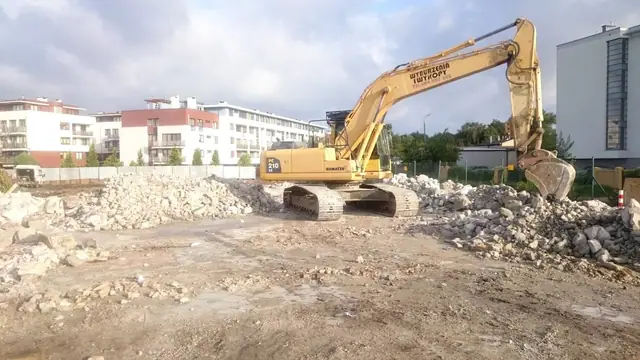 Koparka rozbiera pozostałości ścian, wokół zalegają sterty gruzu betonowego