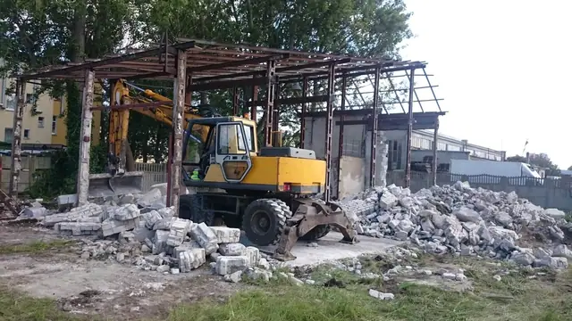 Koparka rozbiera stalowy szkielet hali magazynowej, gruz zalega na ziemi