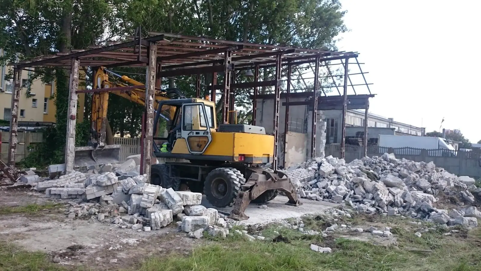 Koparka rozbiera stalowy szkielet hali magazynowej, gruz zalega na ziemi