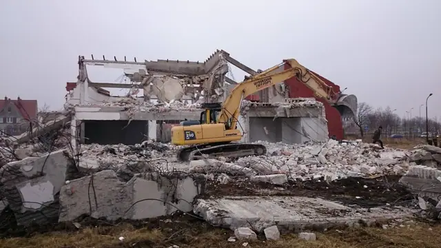 Koparka Komatsu kruszy stropy budynku, przed nią duże bloki betonu z rozbieranej konstrukcji