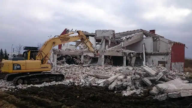 Wyburzanie żelbetowej konstrukcji budynku koparką Komatsu, na pierwszym planie hałdy gruzu betonowego