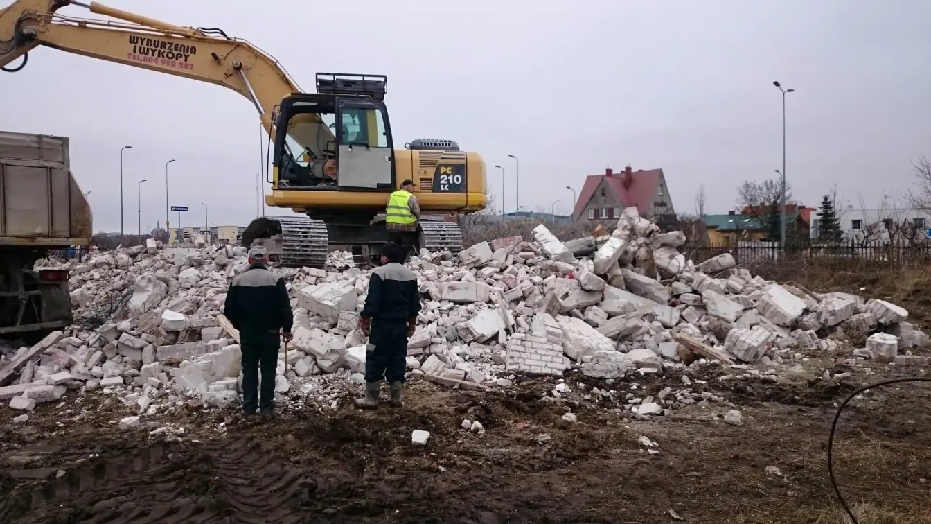 Pracownicy nadzorują załadunek gruzu z rozbiórki, obok koparka Komatsu PC210 i wywrotka
