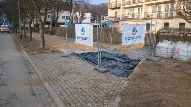 Odgrodzony płotkiem fragment nowego chodnika z kostki brukowej z banerami wykonawcy Szymeta