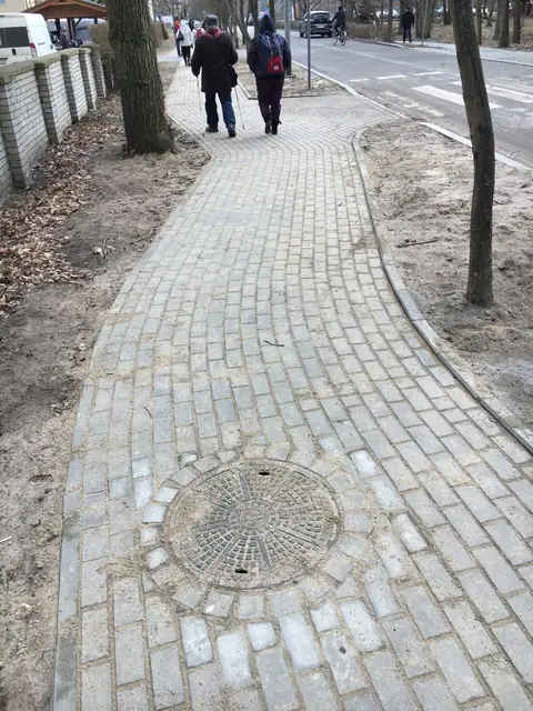 Piesi idą wybrukowaną alejką z kostki granitowej pomiędzy drzewami, widoczny właz kanalizacyjny