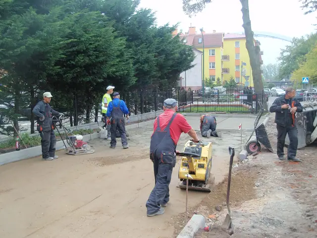 Robotnicy zagęszczają podbudowę zagęszczarką płytową na chodniku przy ogrodzeniu kościoła