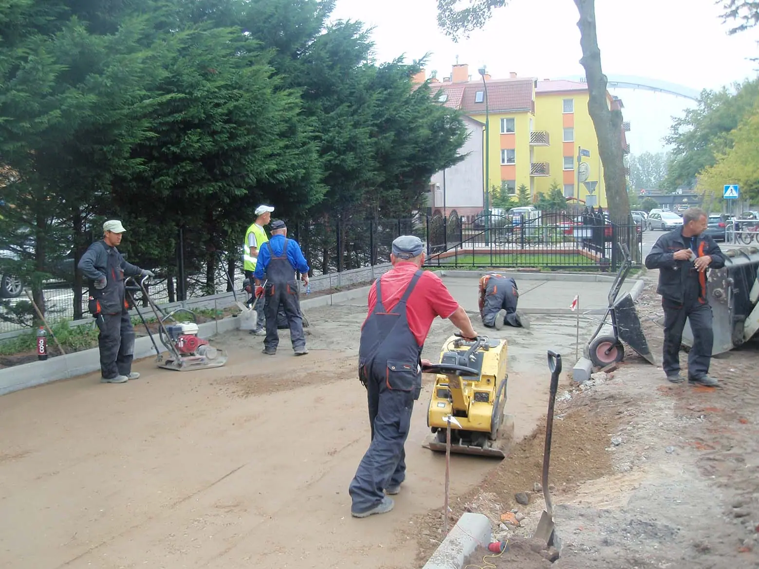 Robotnicy zagęszczają podbudowę zagęszczarką płytową na chodniku przy ogrodzeniu kościoła