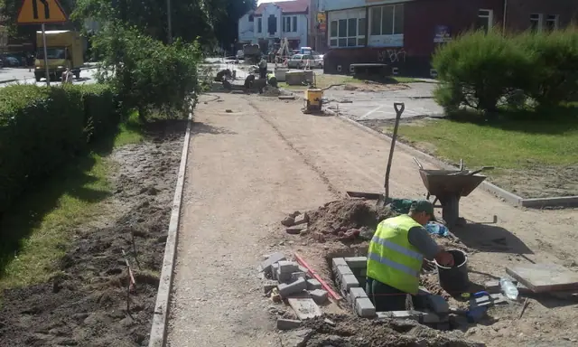 Robotnik układa betonowe obrzeże przy zieleńcu z taczką w trakcie budowy ścieżki rowerowej w Koszalinie