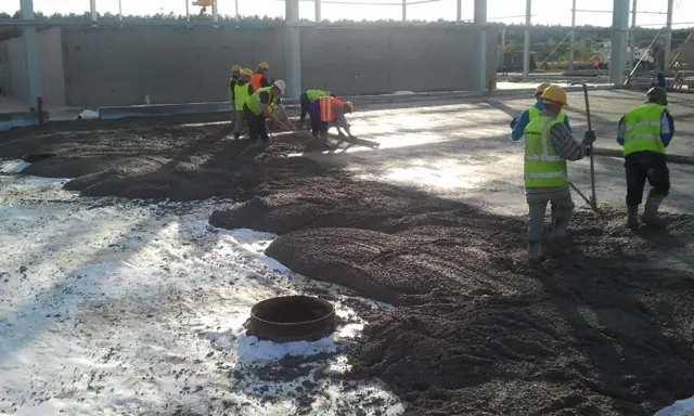 Robotnicy rozprowadzają ciemną warstwę betonu wokół studzienki wewnątrz hali produkcyjnej