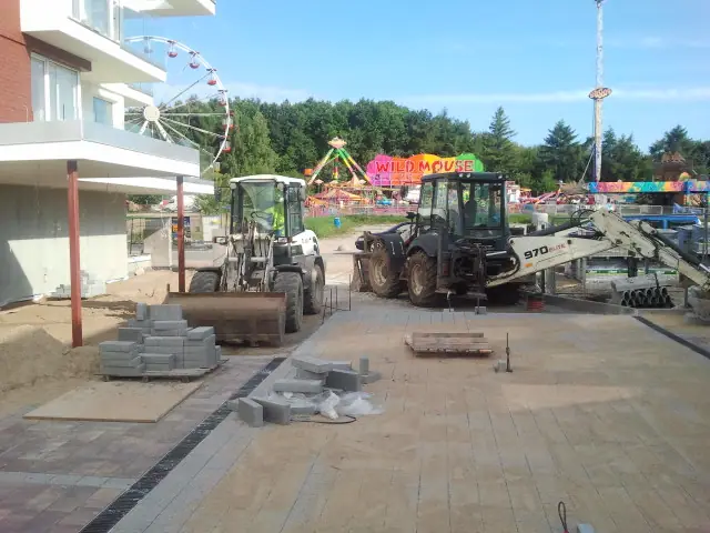 Rozległy wybrukowany plac przed hotelem z zaparkowanymi ładowarkami i atrakcjami w tle