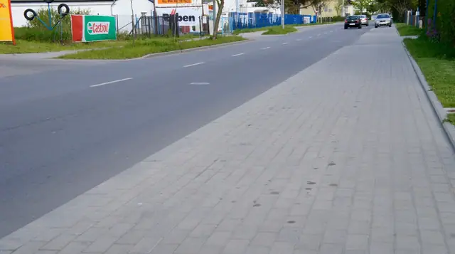 Prosty odcinek wybrukowanego chodnika biegnący wzdłuż gotowej jezdni obok punktów usługowych