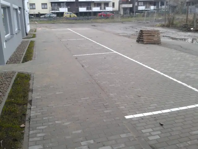 Utwardzony kostką plac z wymalowanymi miejscami parkingowymi, w tle bloki mieszkalne