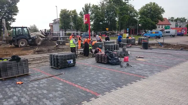Robotnicy układają kostkę brukową na dużym placu, w tle ciągnik i namiot targowy