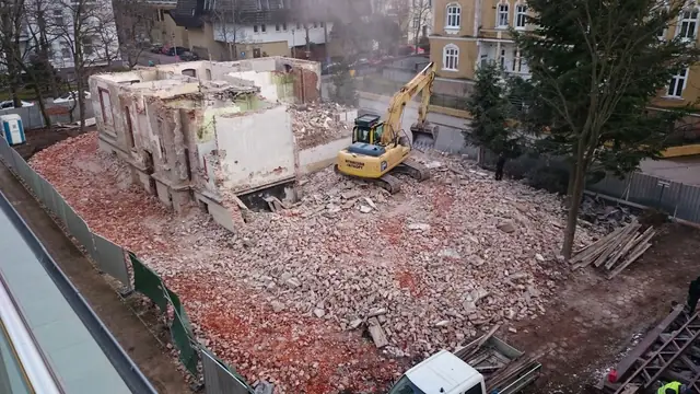 Koparka gąsienicowa wyburza fragment ścian budynku z lotu ptaka, wokół sterty gruzu