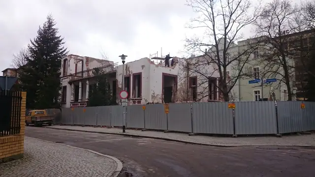 Ogrodzony blaszanym płotem plac rozbiórki widziany od strony skrzyżowania ulic