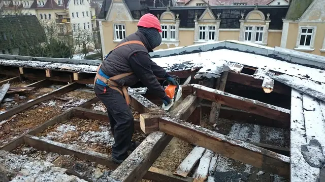 Robotnik w uprzęży piłą spalinową tnie krokwie odsłoniętej więźby dachowej