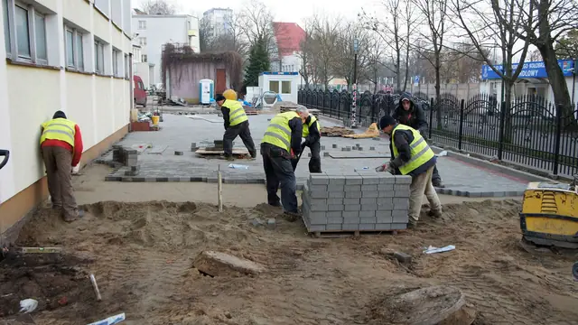Grupa robotników w kamizelkach odblaskowych układa palety kostki brukowej przy elewacji
