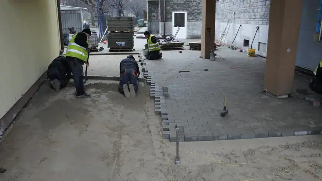 Robotnicy układają kostkę brukową przy wejściu do budynku pod zadaszeniem na słupach
