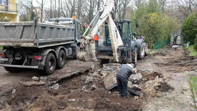 Koparko-ładowarka i ciężarówka usuwają rozbity beton, robotnik zbiera gruz z trawnika