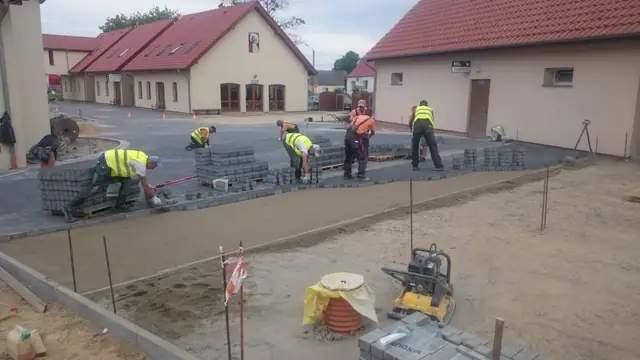 Robotnicy układają kostkę brukową przed budynkiem, obok stoi zagęszczarka płytowa
