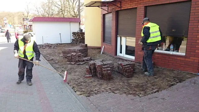 Pracownicy grabią ziemię przy stercie starych cegieł usuniętych spod okien budynku