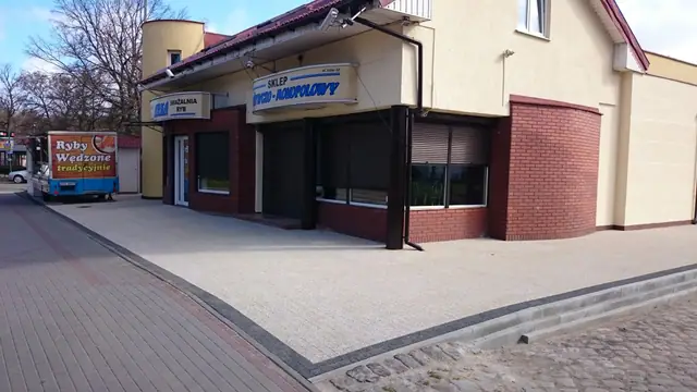 Szeroki plac z płukanego betonu przed wejściem do sklepu, obok widać starą brukowaną ulicę