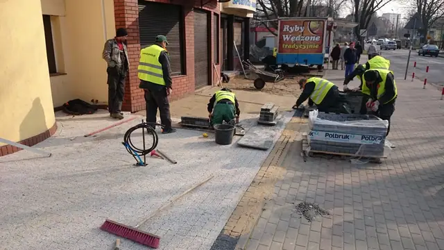 Grupa robotników układa jasną kostkę brukową firmy Polbruk przy użyciu mioteł i palet