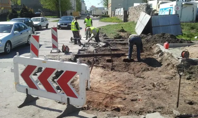Robotnicy pracują przy wykopie na ulicy, osłonięci barierką ostrzegawczą, obok przejeżdżają samochody