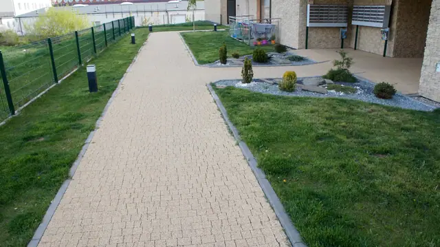 Wybrukowana alejka na trawniku prowadząca do wejścia, otoczona zielonymi krzewami i oświetleniem