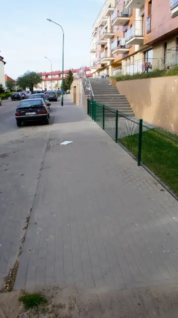 Chodnik z szarej kostki brukowej biegnący wzdłuż zielonego ogrodzenia i skarpy osiedla
